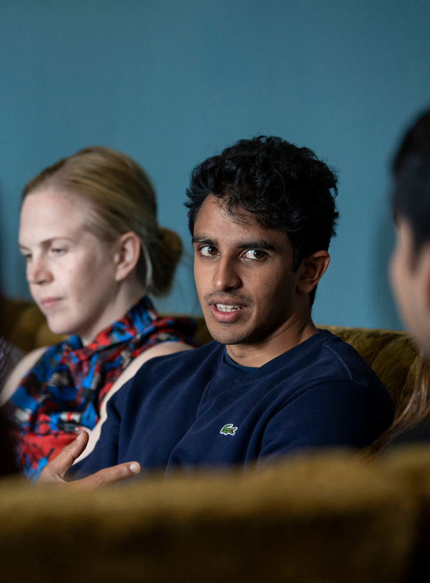 Young man wearing a navy jumper looking toward the camera, his hand raised to emphasise a point he is making in conversation. He is talking to someone out of the camera's line of sight.
