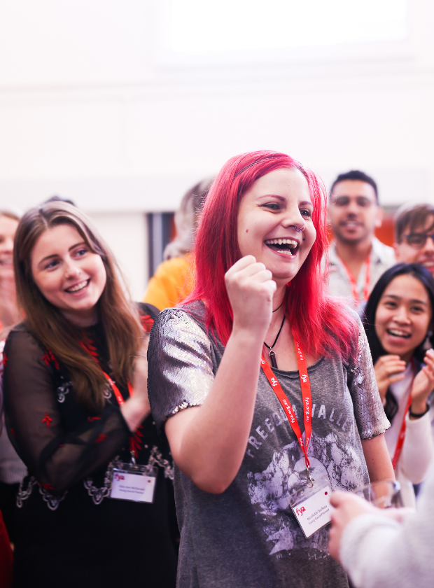 Young woman with red hair wearing silver t-shirt is smiling holding her fist up as part of rock-paper-scissors game. She is surrounded by other young people behind her watching the game play out.