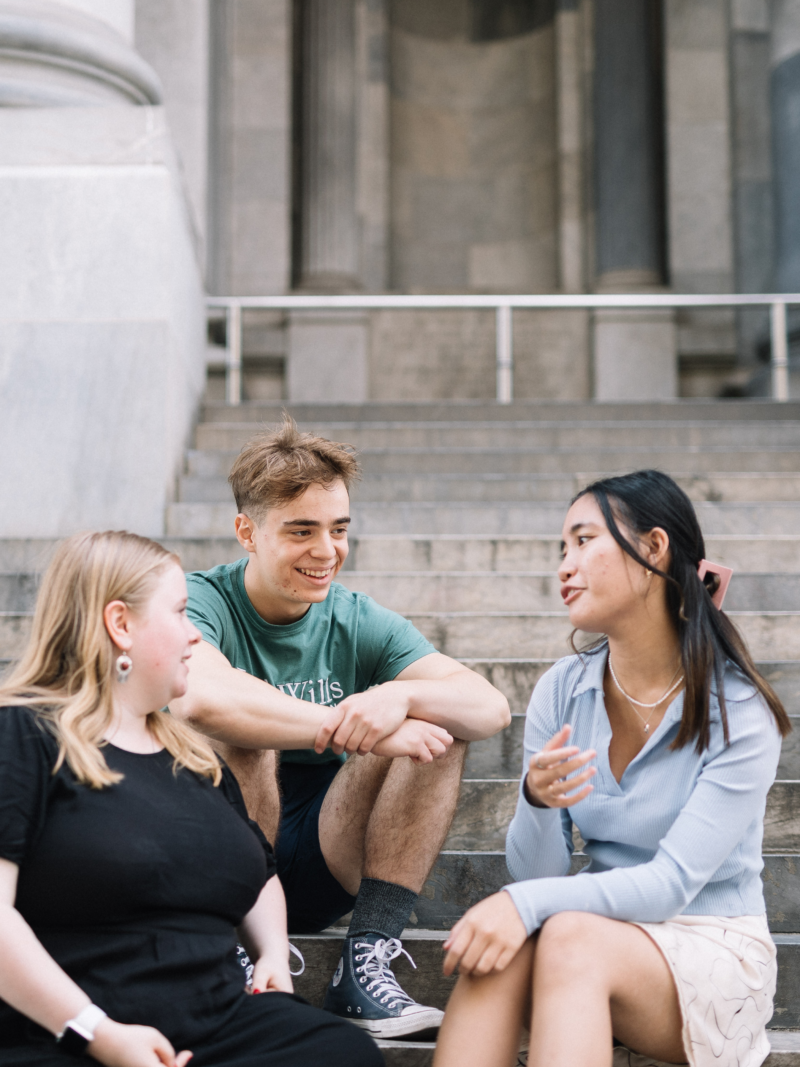 Three young people sit on steps facing one another. The person on the right wearing a blue long sleeve top with long dark hair is speaking.