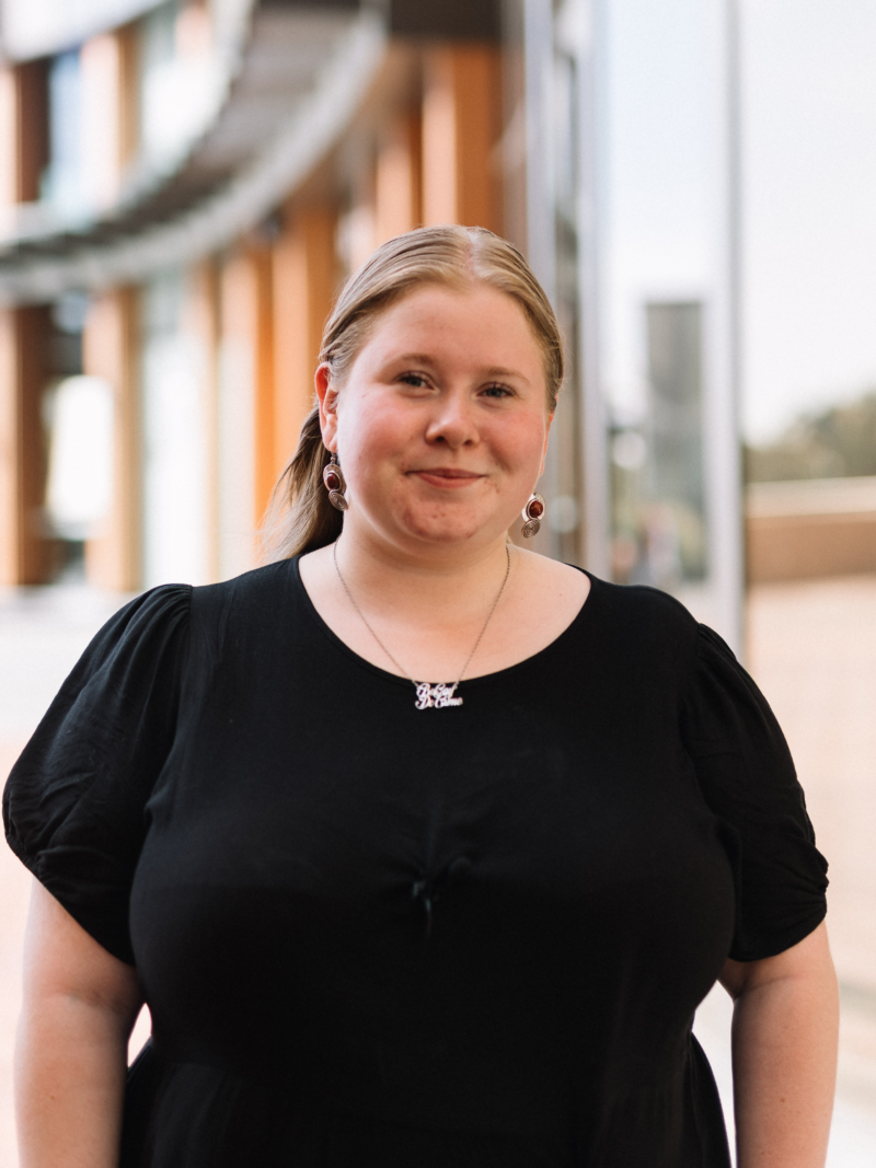 Tabitha is wearing a black short sleeve top, with her long blonde hair behind her back, and is smiling at the camera.